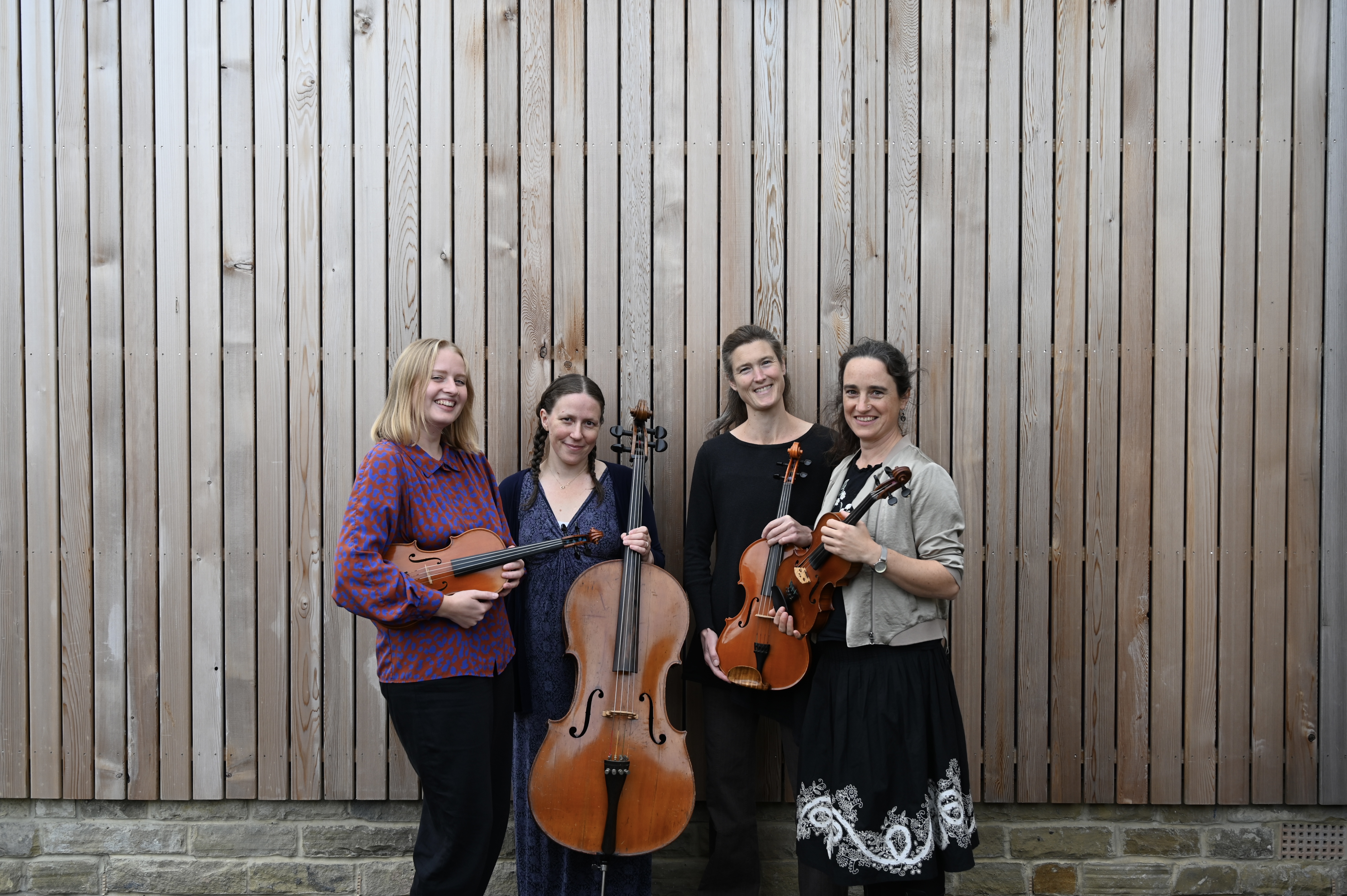 Four musicians stand against a wood building, holding string instruments and looking at each other.